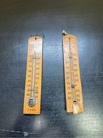 Two wooden advertising thermometers side by side on a dark surface showing full length and condition.