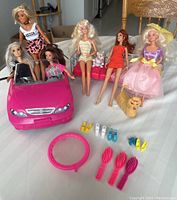 Full view of 6 Barbie dolls including 4 seated, 2 standing, with pink convertible car in front, pink couch with cat figure, dog figure, trampoline and accessories laid out on white tablecloth.