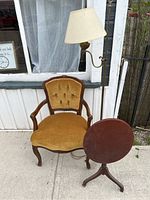 Three-piece set of chair, folding side table, and floor lamp shown together outside a building. Chair is wooden with golden upholstery, lamp is brass with fabric shade, table is round wooden folding table.