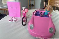 Pink Barbie car with seated Barbie doll, pink bicycle with helmet, pink and white small shoulder bag, and closed pink armoire on white surface with window light.