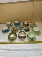 Photo of ten assorted porcelain cups and saucers arranged on a white cloth, showing various colors including blue, green, white, and cream bases with floral decorations and gold trim.