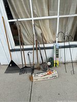 Photo showing multiple rakes, shovels, hoes, garden stakes, a boxed hedge trimmer, and the rain watering wand in packaging arranged against a window.