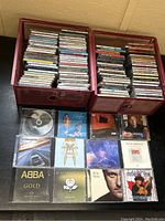Two red crates filled with over 50 music CDs stacked inside. Crates placed on floor against wall.