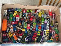 Wide view of a cardboard box filled with over 90 loose Hot Wheels cars in various colors and styles including muscle cars, sports cars, and fantasy vehicles.