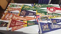 Photo showing a large assortment of colorful vintage felt pennants laid out on a table, from various locations in Canada and USA such as Niagara Falls, Festival of Britain 1951, Banff Chair Lift Mt. Norquay, Cape Breton, and Medicine Hat.