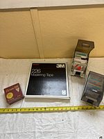 Photo showing sealed Dysan 100 diskette box, 3M 226 Mastering Tape box, and two plastic holders with floppy disks on white cloth by a wall.