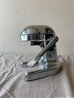 Front angle view of retro mid-century modern chrome manual juicer on cloth surface with neutral wall background.