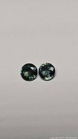 Pair of round multicoloured sapphires on white background showing green and blue hues, used for detailed visual assessment of color and cut.