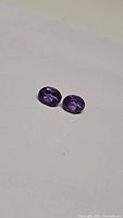 Two oval cut purple amethyst gemstones lying on a white surface, showcasing their color and faceted cut.