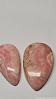 Two natural rhodochrosite pear-shaped cabochons laid side by side showing their polished, glossy surfaces and characteristic pink and cream color patterns with brown inclusions.