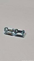 Two emerald cut blue topaz gemstones, approximately 6x4mm each, shown on gray background.