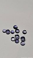 Top view of ten small round cabochon cut purple iolite gemstones on a neutral background showing their size and color.