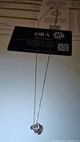Full view of the silver chain with heart-shaped pendant featuring a small moissanite gemstone and associated GRA certificate card visible in the background.