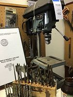Full view of Delta drill press with assorted drill bits in front, showing the drill press head, table, and mounted drill bits, dusty from storage.