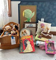 Wide shot of wooden doll crib with plush bears, fabric art, framed embroidery, plate, paper dolls book, baby blankets, basket, and boxed dolls.