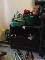 Garden tools and accessories on and around a dark wooden bench, including green plastic tub and red funnel