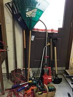 Image showing collection of garden rakes, shovel, electric trimmer with cord, hand tools in yellow plastic basket, gas weed trimmer, pitch fork, and spades, positioned against garage door.