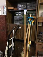 Full view of dark wood shelving unit with plastic hardware storage drawers filled with assorted nuts, bolts and washers. Tools leaning in front include snow shovels, and push broom.