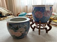 Two large blue koi pots with pink floral designs; one pot on wooden stand and other on floor.