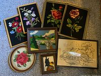 Eight framed needlepoint and punch yarn art pieces arranged on carpet, showing floral and landscape designs