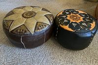 Two round ottomans on carpet with visible wear; smaller black and tan with tribal embroidery, larger beige and brown with star pattern and scenes