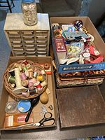 Full view of sewing baskets with spools of threads, buttons, scissors, a thimble, and a hand mirror on a table with some boxes.