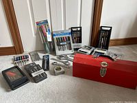 Wide view of assorted tools laid out on carpet including socket sets, screwdriver sets, adjustable wrench, tape measure, and red metal toolbox.