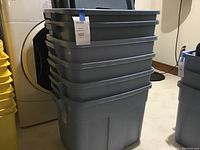 Stack of three large gray Rubbermaid plastic storage totes with lids, tagged for sale, in basement laundry room.