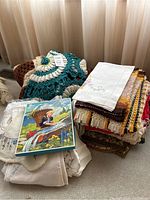 Lot of assorted folded crochet tablecloths, napkins, placemats, and some decorative textiles stacked together with a vintage print textile on top