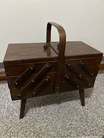 Closed wooden sewing box with handle, showing exterior wooden finish and legs.
