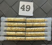 Top view showing 10 glass vials filled with gold flakes placed side by side on a dark surface with a lot number 49.