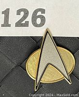 Front view of Star Trek communicator badge pin showing gold oval background and silver arrowhead shape with black outline