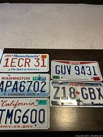 Five American license plates laid out on brown surface with beige carpet background, showing distinct states and numbers