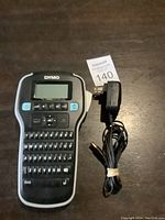 Dymo D1 label printer and charger placed on a wooden surface, including MaxSold lot tag 140
