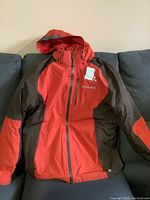 Photo of medium Columbia winter jacket in red and dark grey, laid flat on a couch showing front with hood and zipper closed.