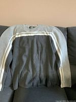 Full view of men's Roots sweater laid flat on couch showing front side with dark grey body and light grey sleeves with white stripes across chest and arms.