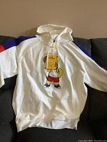 Full front view of white hoodie laid flat showing the large embroidery of a cartoon character with a yellow head resembling a basketball and wearing shorts and shoes.