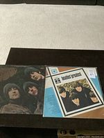 Two Beatles vinyl LP records placed side by side on a dark wooden surface. Left is Rubber Soul album cover showing band members illustration, right is Beatles' Greatest compilation in sealed packaging.