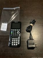 Photo showing TI-84 Plus CE calculator and power supply charger on a dark wooden surface with plastic bag and auction tag