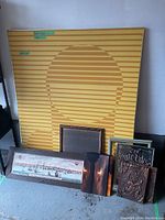 Collection of miscellaneous framed wall art leaning against a wall and on the floor, including a large abstract piece with yellow and orange stripes, several smaller framed pieces including a cityscape, a relief panel, and vintage text art.