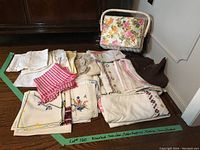 Assorted linen items arranged on floor including napkins, doilies, dish cloths, and a floral sewing basket.