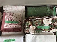 Photo showing folded and stacked assorted table linens including floral patterned tablecloths, solid green fabric, and pink patterned squares with labels visible.