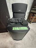 Lot includes one Bose Acoustimass subwoofer module, two small Bose speakers stacked on top, power cords, and a remote control placed on top of the subwoofer module.