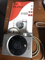 Silver Canon ELPH Ixus film camera with matching user manual on wooden surface