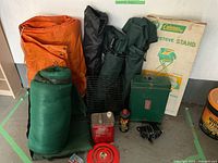 Camping gear including green sleeping bag, orange tarp, Coleman stove with stand in box, folding camp chairs in bags, campfire grill and fuel cans shown on floor against white wall.