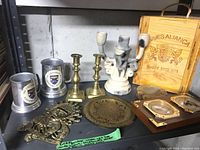 Vintage wooden wine box with etched design on front, brass plaques including lion heads and floral plate, brass candle holders, ram head candle holder, and two pewter University of Western Ontario beer steins on metal shelving.