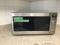 Front view of Panasonic microwave showing stainless steel body and black glass door. Digital display on top right shows time 2:39. Green label with lot number taped on front.