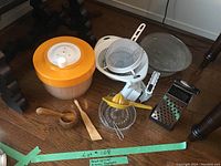 Photo of the entire lot showing an orange and beige salad spinner, plastic colanders, metal strainers, various graters, citrus juicers, and wooden kitchen tools.