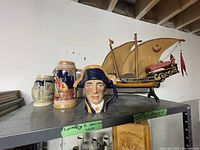 Three vintage beer steins and a handmade model ship displayed on a metal shelf in a basement setting.