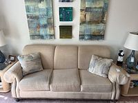 Front view of beige upholstered couch with three seat cushions, rolled arms, nailhead trim, two patterned throw pillows, and wooden bun feet. Neutral tone with matching living room decor around.